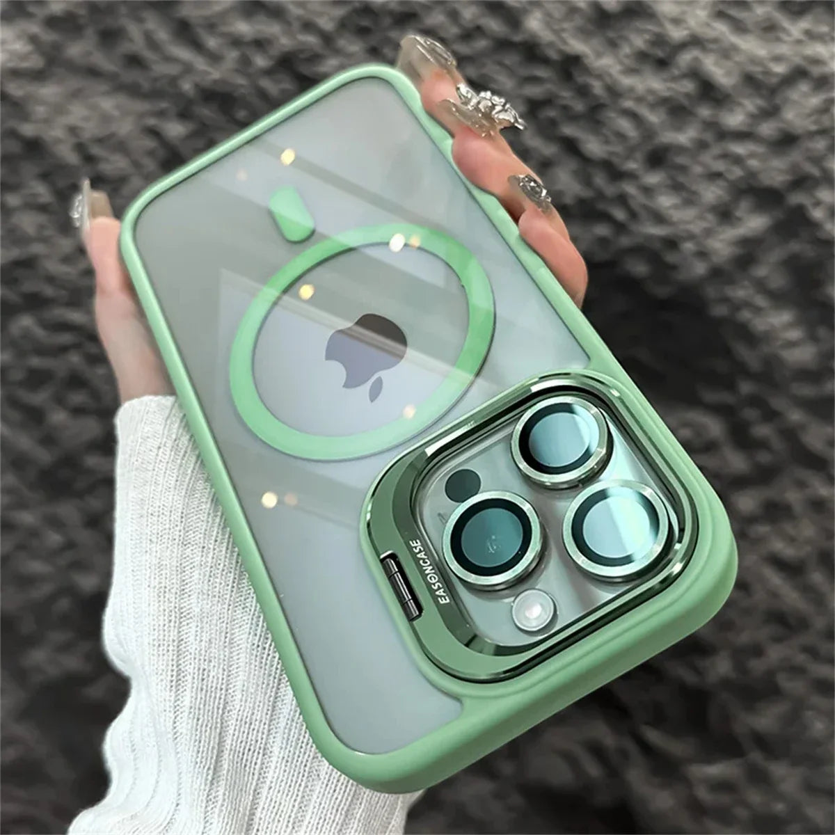 A hand with long, decorated nails holds a smartphone with a green, translucent case. The phone features three camera lenses, and the iconic bitten apple logo is visible on the back.