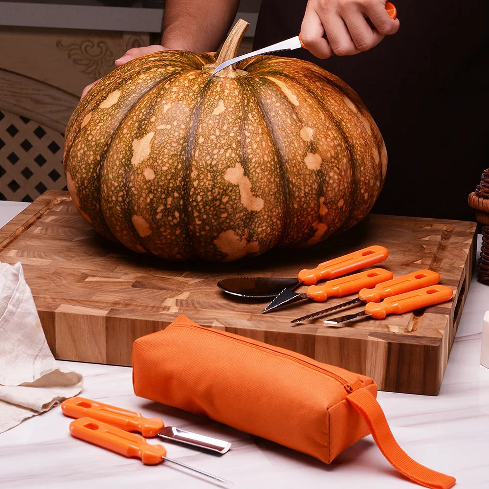 Person carving a large pumpkin on a wooden board with a serrated knife. Various orange-handled carving tools and an orange pouch are laid out on a marble surface nearby.