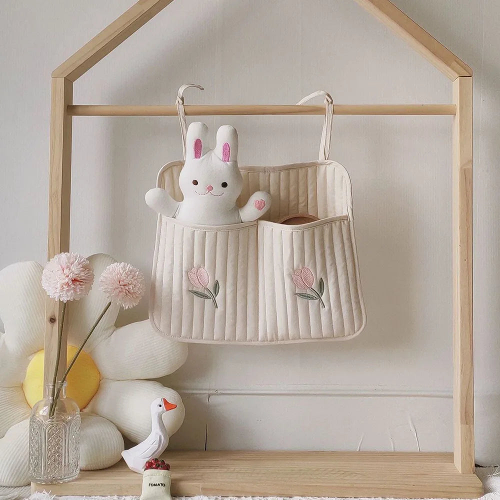 A cozy scene with a plush bunny in a hanging fabric organizer, embellished with pink flowers. Nearby, pink flowers in a vase and a white goose figurine sit on a wooden shelf.
