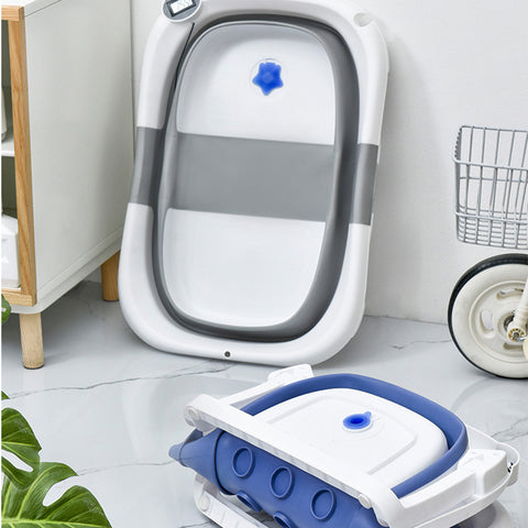 Foldable baby bath tubs in gray and blue are neatly arranged on a tiled floor. They are designed for compact storage, placed near a small wooden cabinet and a laundry cart.