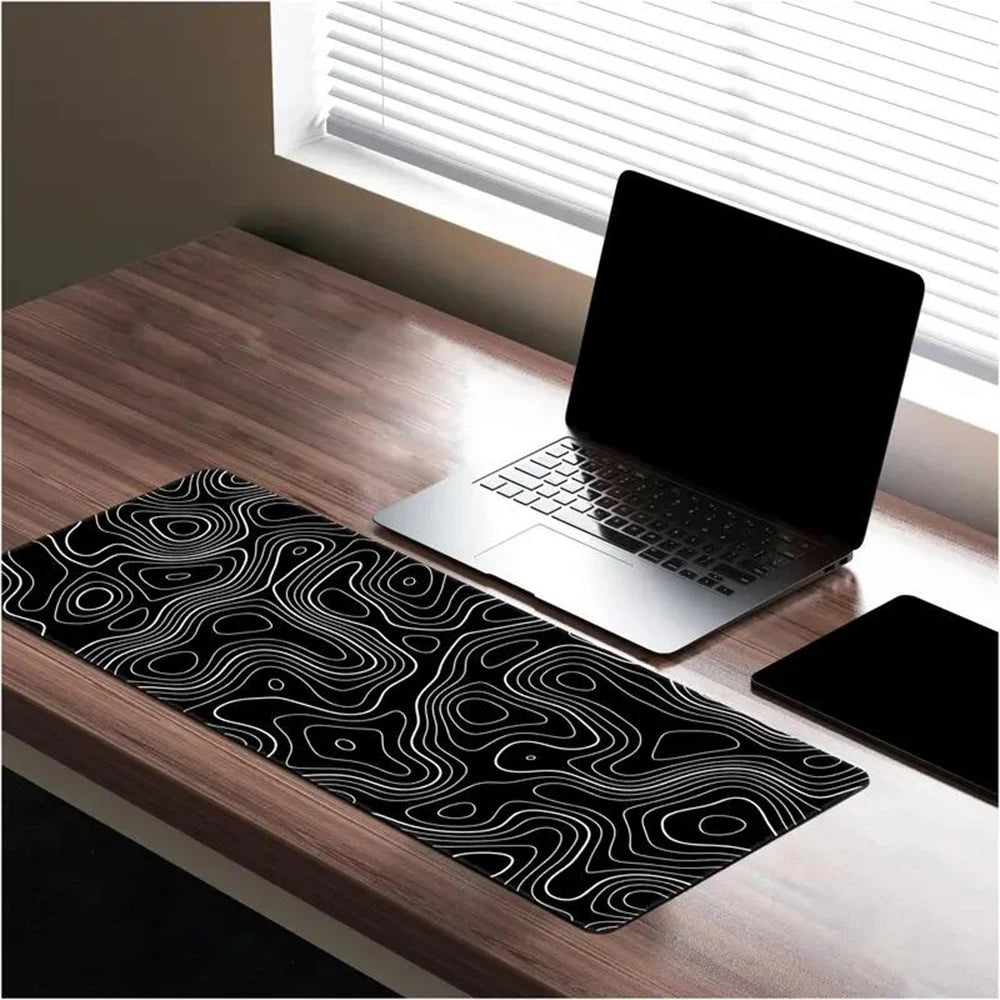 A laptop on a wooden desk near a window with blinds. The desk features a black desk mat with a white abstract line pattern, and a closed tablet lies beside the laptop.