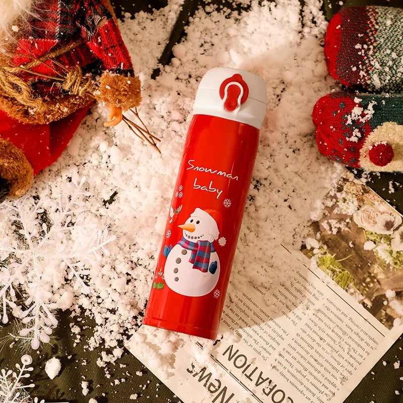 Red thermos with a snowman design, labeled "Snowman baby," on snowy ground. Surrounding items include a knitted glove, Santa toy, snowflake, and a piece of paper.