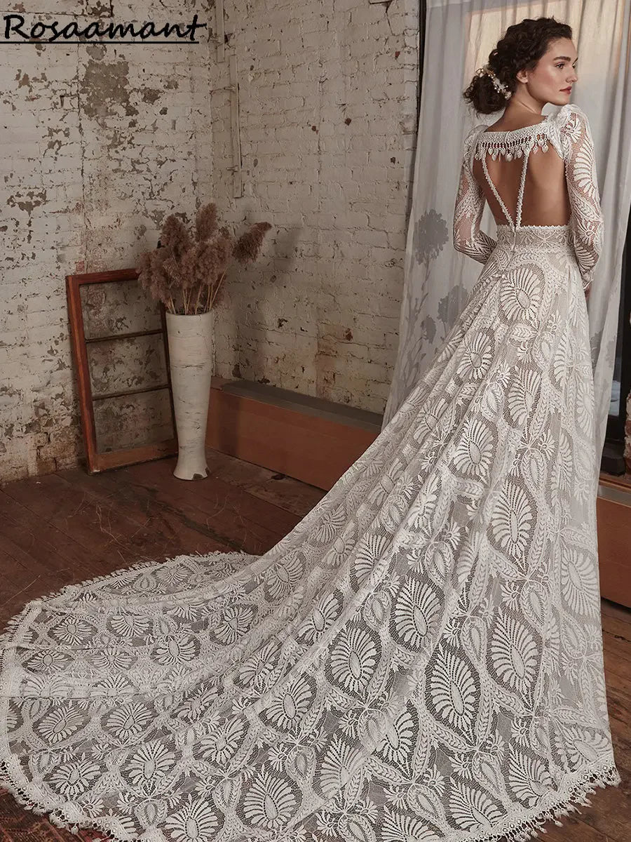 Woman in a lace bridal gown with an intricately designed train stands in an industrial-style room with exposed brick walls and dried flowers in a vase.
