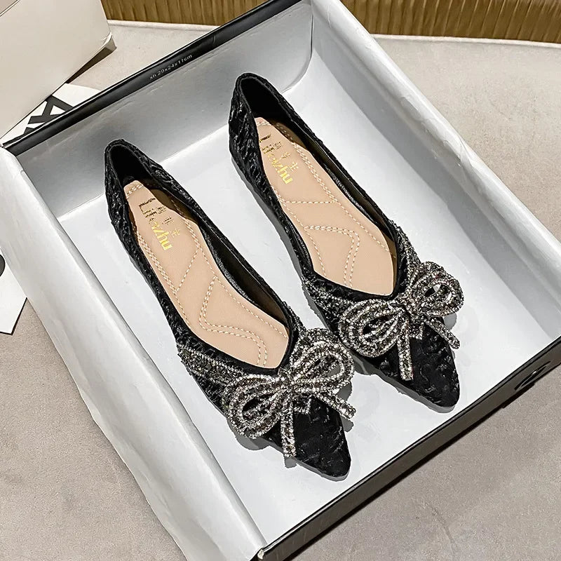 Black velvet flats with decorative silver bows and rhinestone details, displayed in an open shoebox on a soft beige surface.