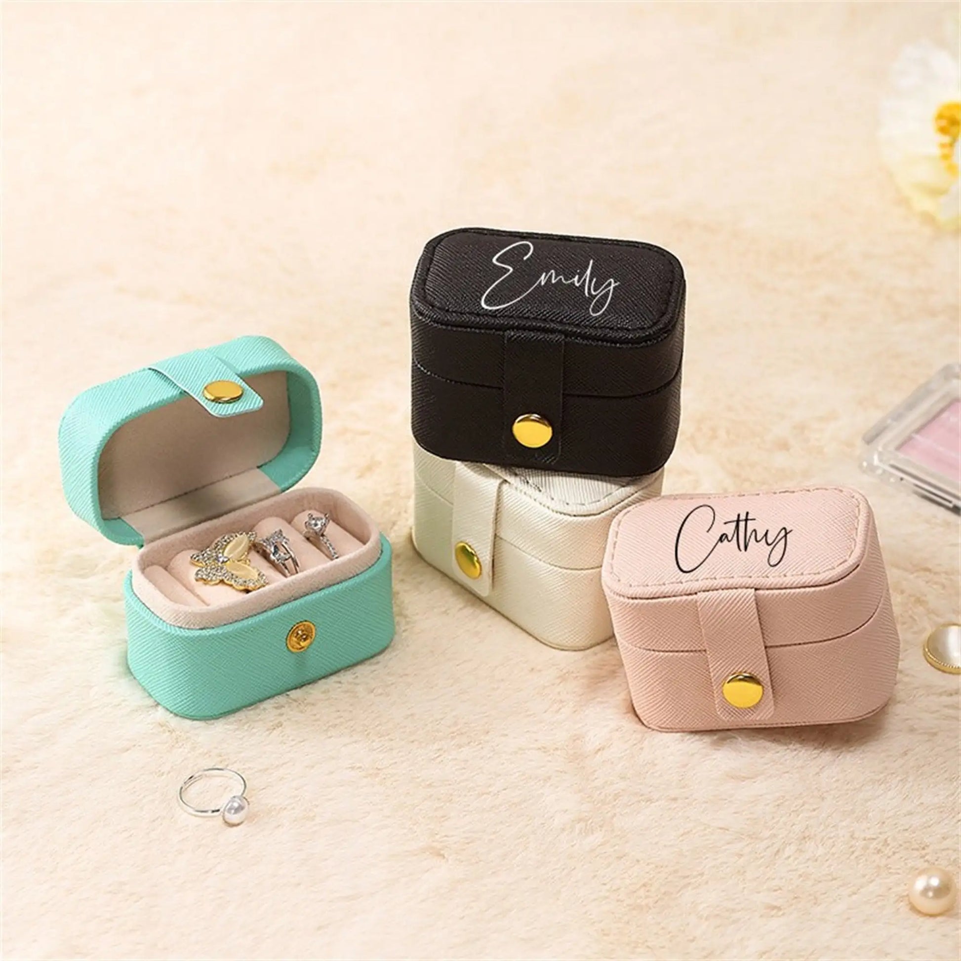 Stacked jewelry boxes in teal, black, cream, and pink, some with names, on a soft surface. The teal box is open, displaying rings and a necklace. Loose pearl ring nearby.
