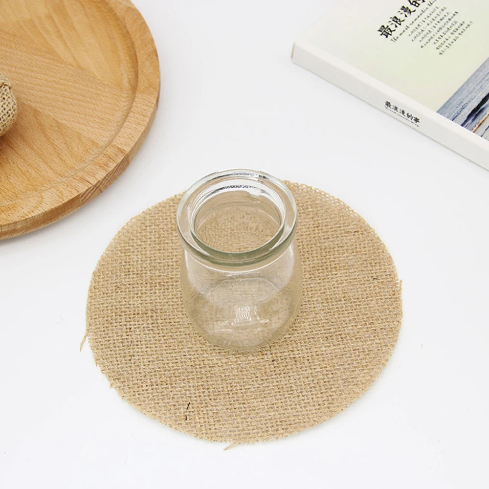 A small glass jar sits on a round burlap mat, next to a wooden tray and an open book with text on a white surface.