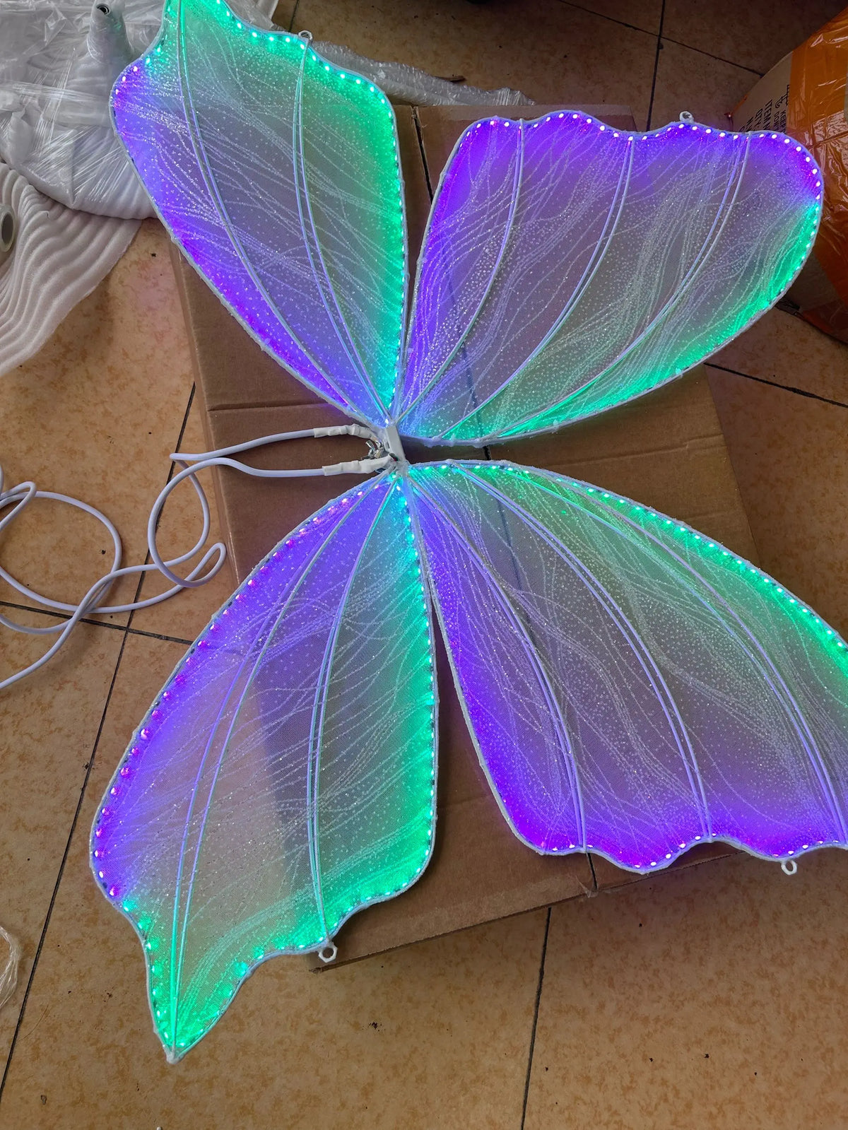 Brightly lit butterfly wings with green and purple LED lights, displayed on a cardboard surface. Loose wires are visible, suggesting they are not yet installed.