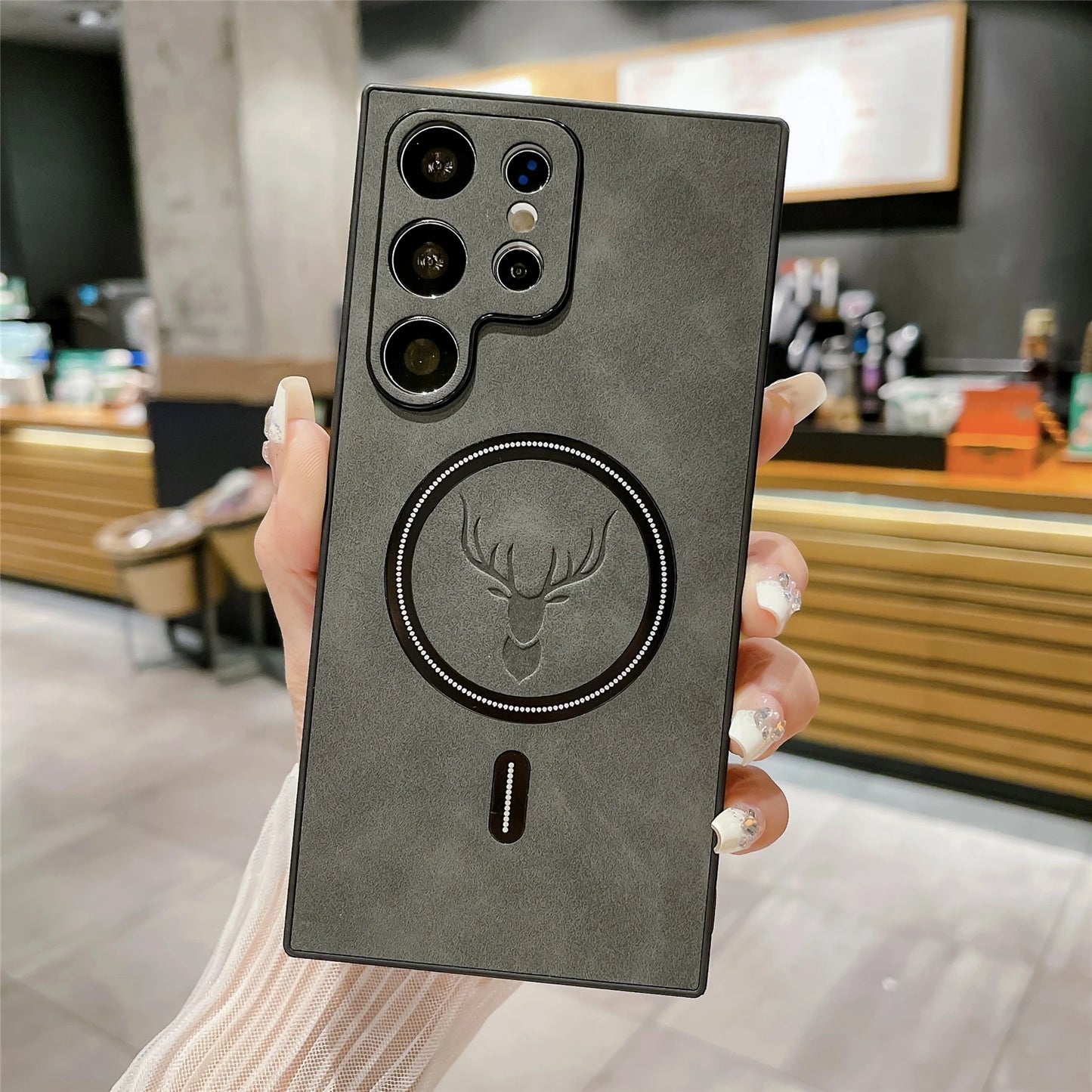 A hand holds a smartphone with a multi-camera setup and a leather-style case featuring an embossed deer logo. The background shows a blurred indoor setting with wooden elements.