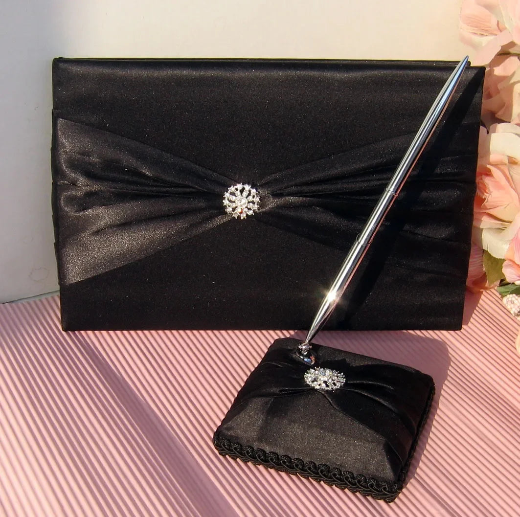 Elegant black guest book with a satin finish, adorned with a sparkling brooch. Paired with a matching pen set on a decorative cushion, placed on a striped surface.
