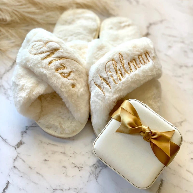 Fluffy white slippers embroidered with "Mrs." and "Willman" in gold, paired with a white gift box tied with a gold ribbon, all set on a marble surface.