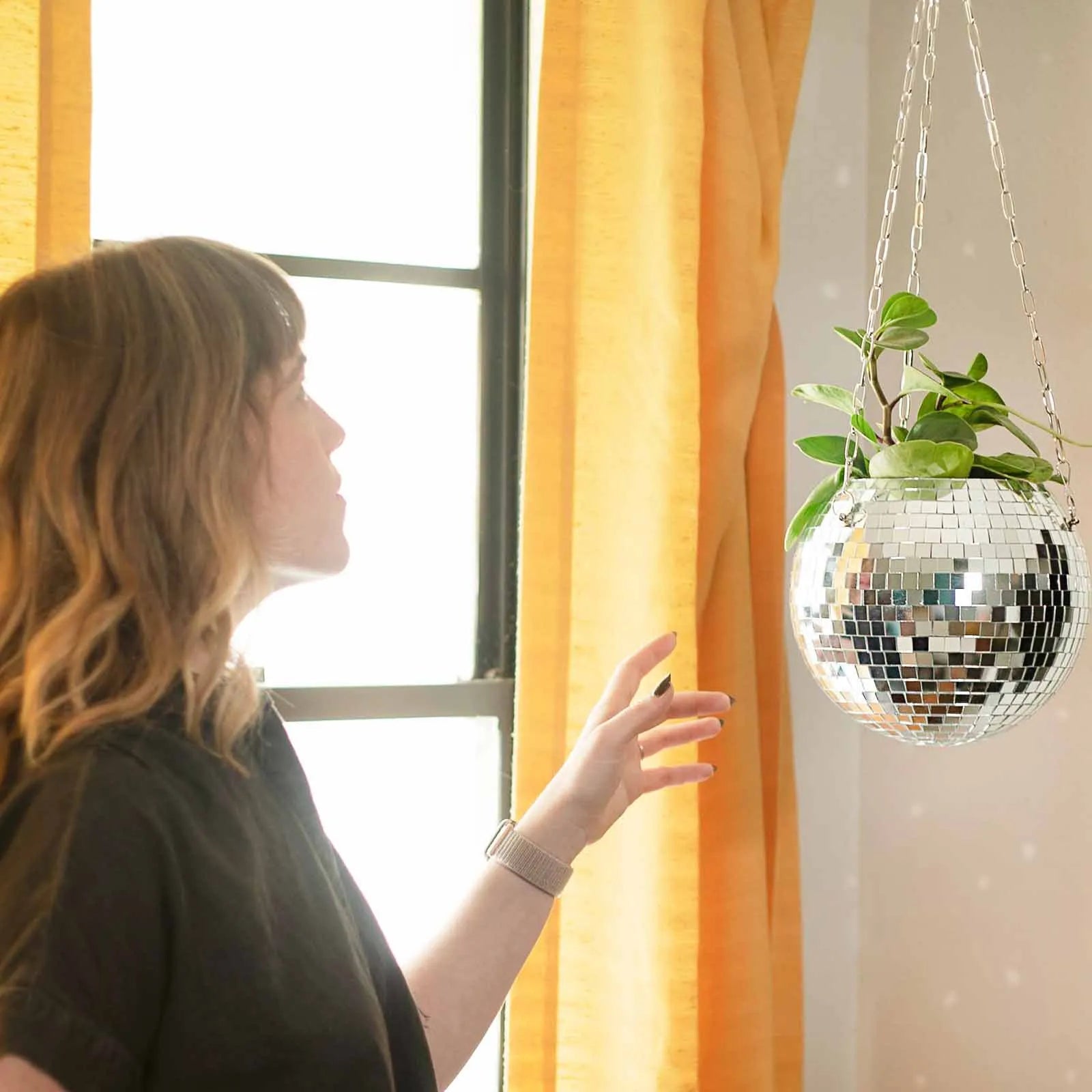 A person in a black shirt reaches toward a hanging disco ball planter with green leaves, next to a window with yellow curtains, creating a vibrant indoor scene.
