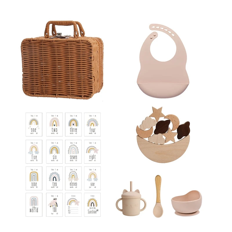 A wicker basket, a pink silicone bib, wooden toys in a bowl, milestone cards, an ear-shaped sippy cup, a wooden spoon, and a pink suction bowl are displayed against a white background.