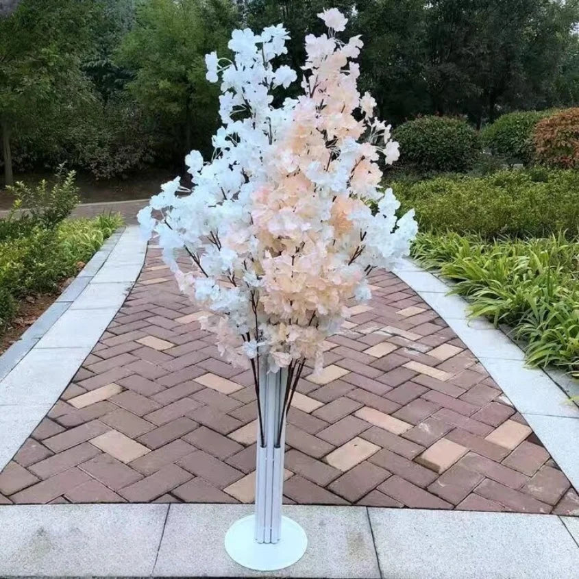 Artificial cherry blossom branches in pastel pink and white are arranged in a tall white stand on a brick pathway, surrounded by lush greenery in a garden setting.