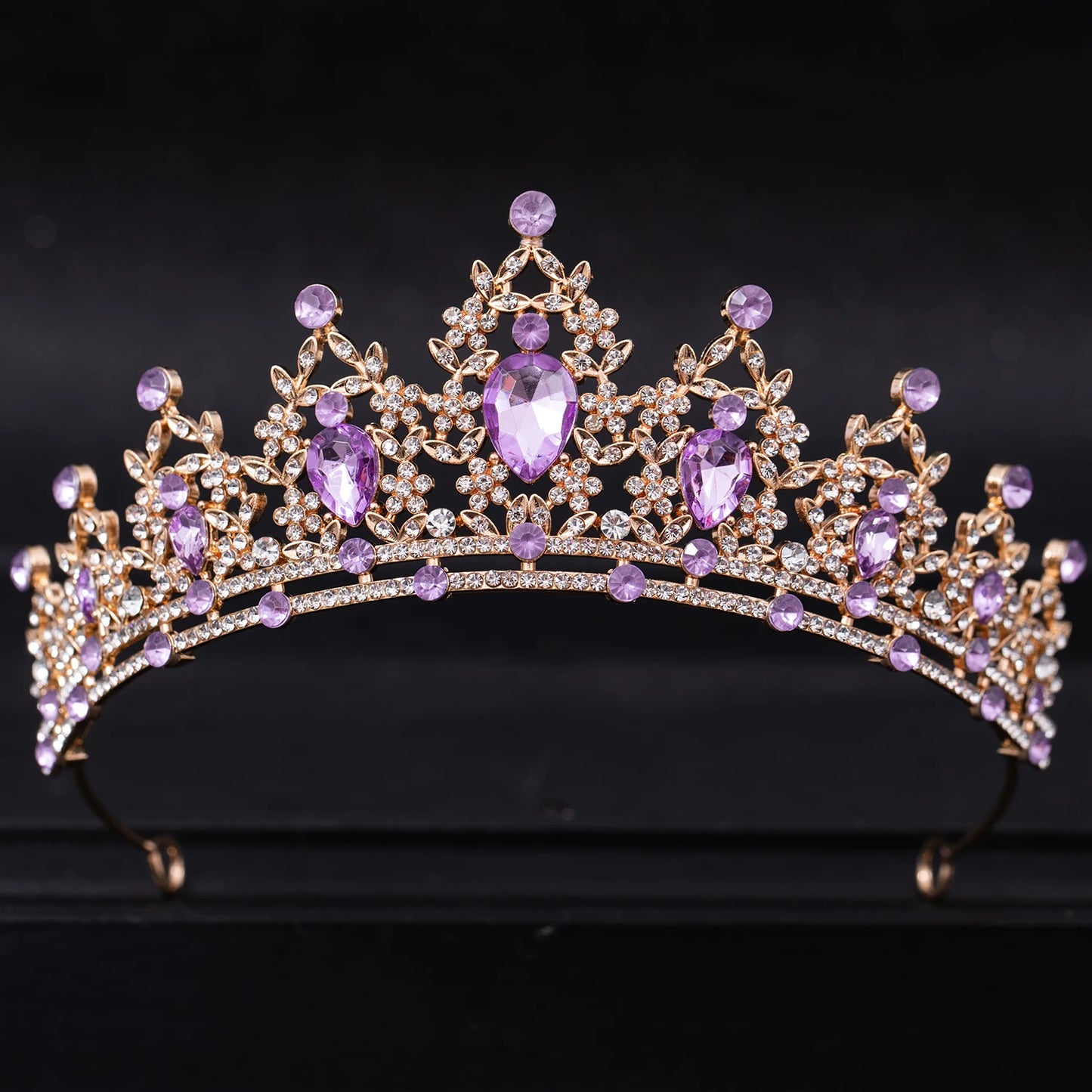 Elegant gold tiara adorned with purple teardrop and round crystals, intricately designed with smaller sparkling clear stones, set against a dark background.