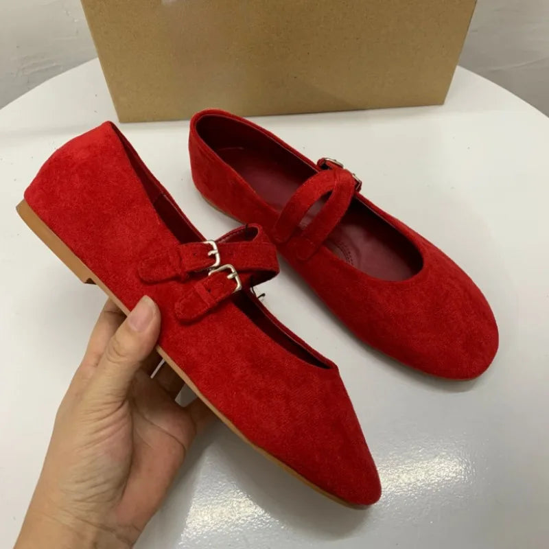 A pair of vibrant red suede ballet flats with dual buckle straps; one shoe is held in a hand, and the other rests on a white surface against a plain cardboard background.