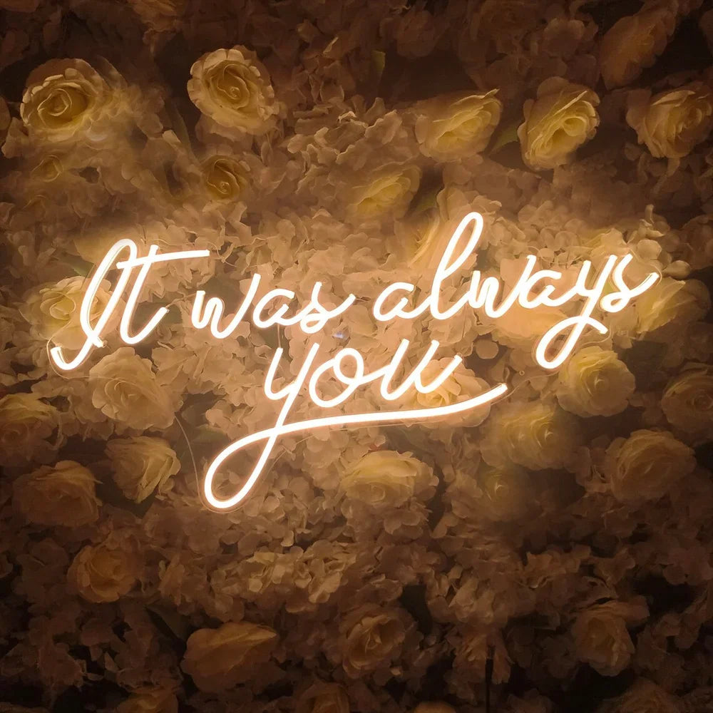 Neon sign with cursive text "It was always you" glowing against a backdrop of white roses, creating a romantic and warm ambiance.