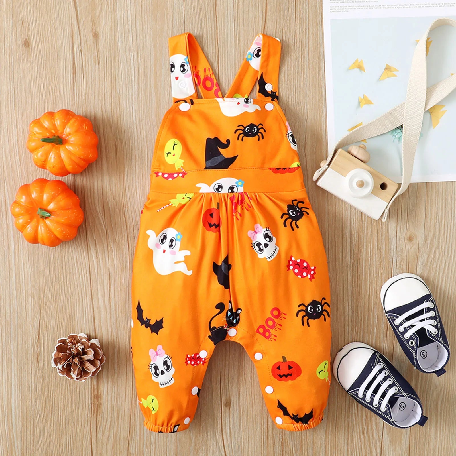 Orange Halloween-themed baby romper with ghosts and pumpkin designs, surrounded by two mini pumpkins, pinecone, toy camera, and black-and-white baby sneakers on wood floor.