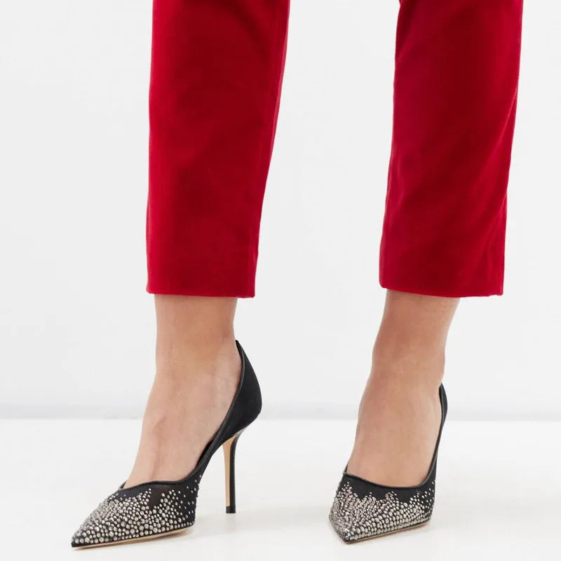 Woman wearing red pants and black stiletto heels with sparkling rhinestone embellishments, standing on a light-colored surface.