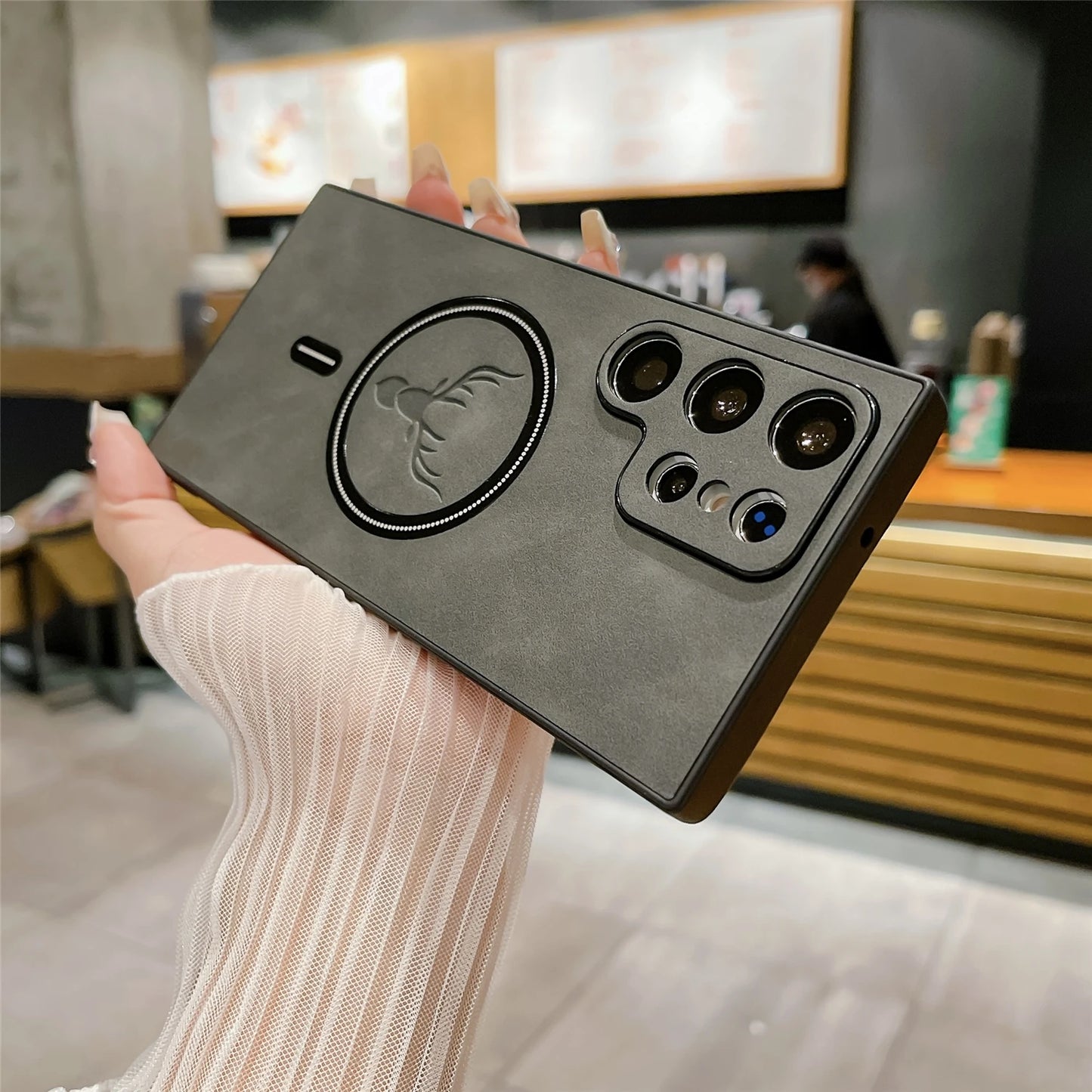 Hand holding a smartphone with a black case, featuring a circular design and multiple rear cameras. Background shows a blurred wooden and concrete café interior.