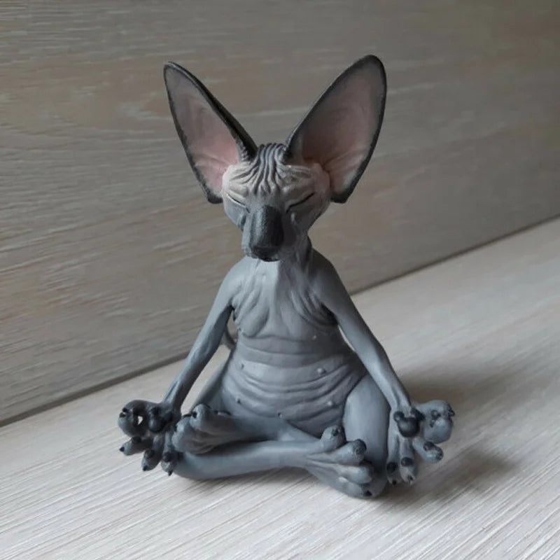 A figurine of a hairless cat with large ears in a meditative pose, seated cross-legged on a wooden surface. The cat's eyes are closed, and its front paws rest on its knees.