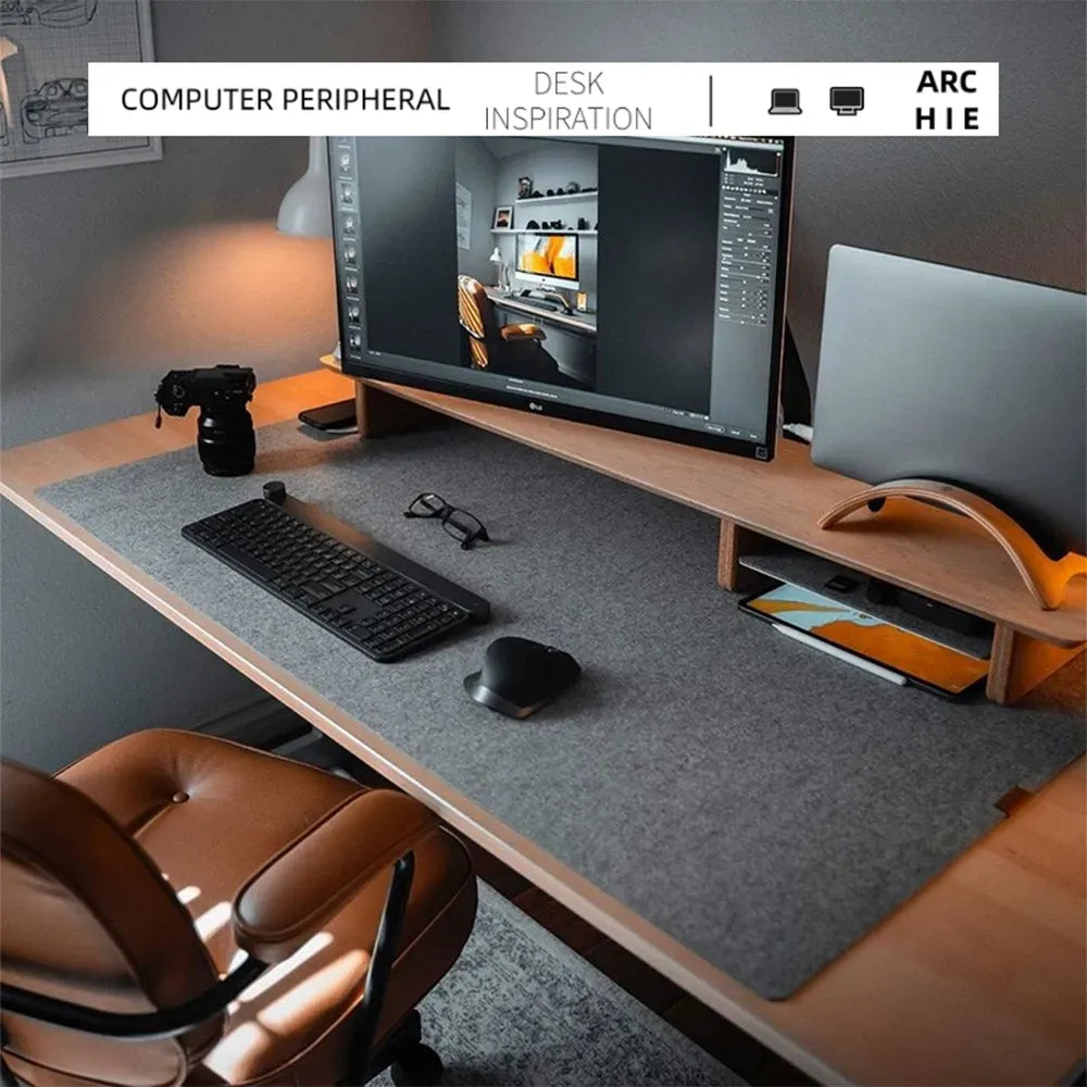 Modern desk setup featuring a large monitor, keyboard, mouse, camera, and laptop stand on a wooden desk with a gray mat. An orange-brown chair completes the workspace.