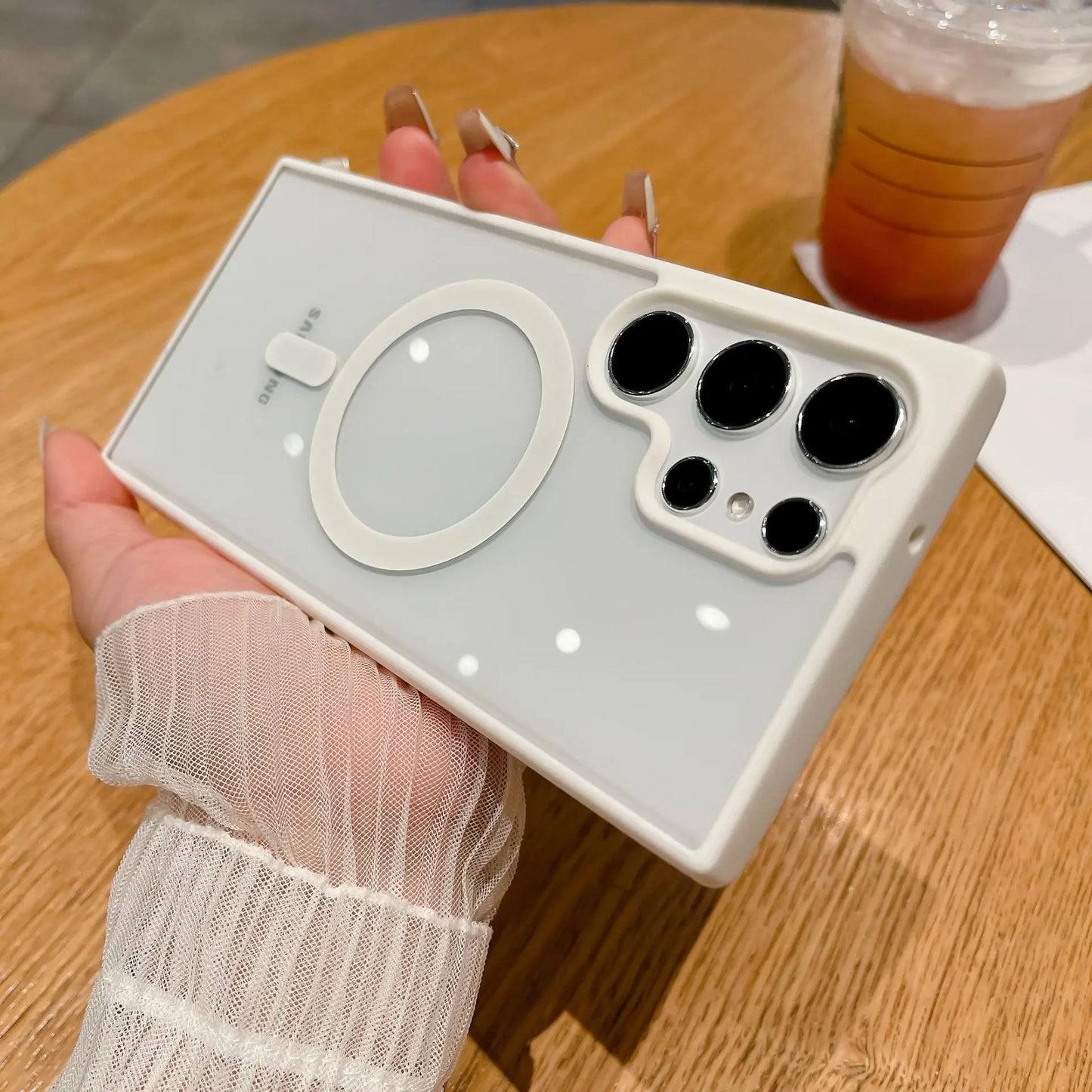 A person holds a smartphone with a distinctive white case featuring a circular design and multiple lens cutouts. A drink in a plastic cup is in the background on a wooden table.