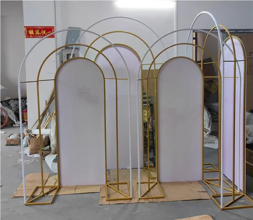 Decorative gold and white arch frames with white panels, arranged in a cluttered workshop. Cardboard sheets are placed on the ground beneath the arches.