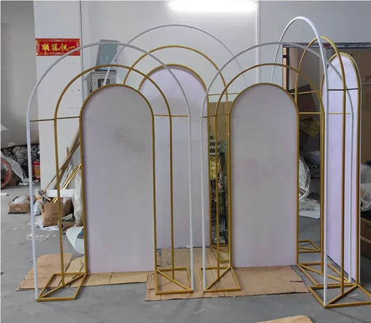 Decorative gold and white arch frames with white panels, arranged in a cluttered workshop. Cardboard sheets are placed on the ground beneath the arches.