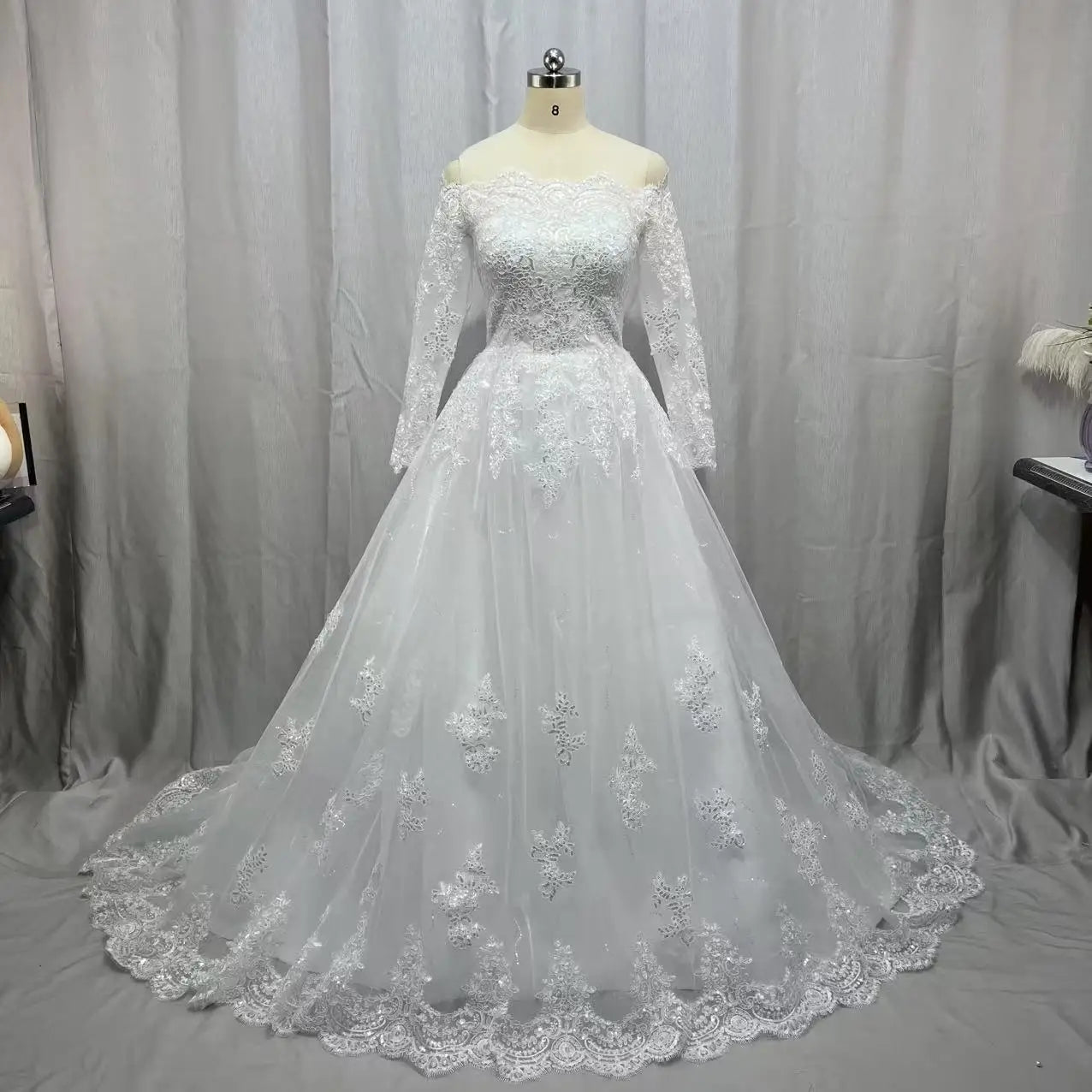 A white lace wedding dress on a mannequin with off-the-shoulder long sleeves and a flowing skirt adorned with intricate floral patterns, set against a soft gray curtain backdrop.