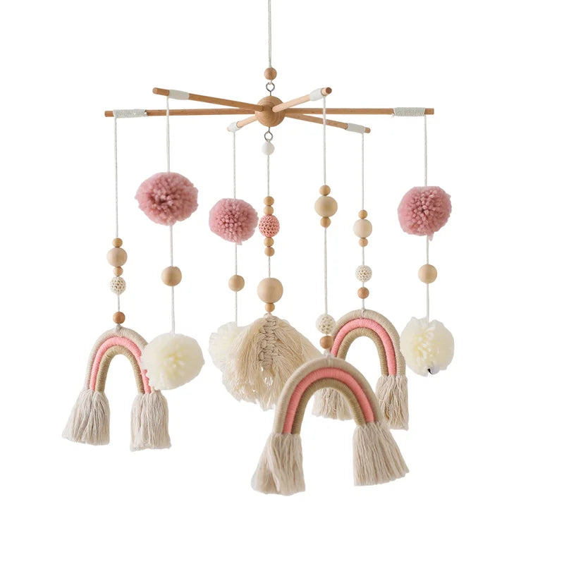 Hanging baby mobile with wooden frame, featuring pink and cream yarn pom-poms, woven rainbows, and wooden beads, creating a soft and whimsical design.