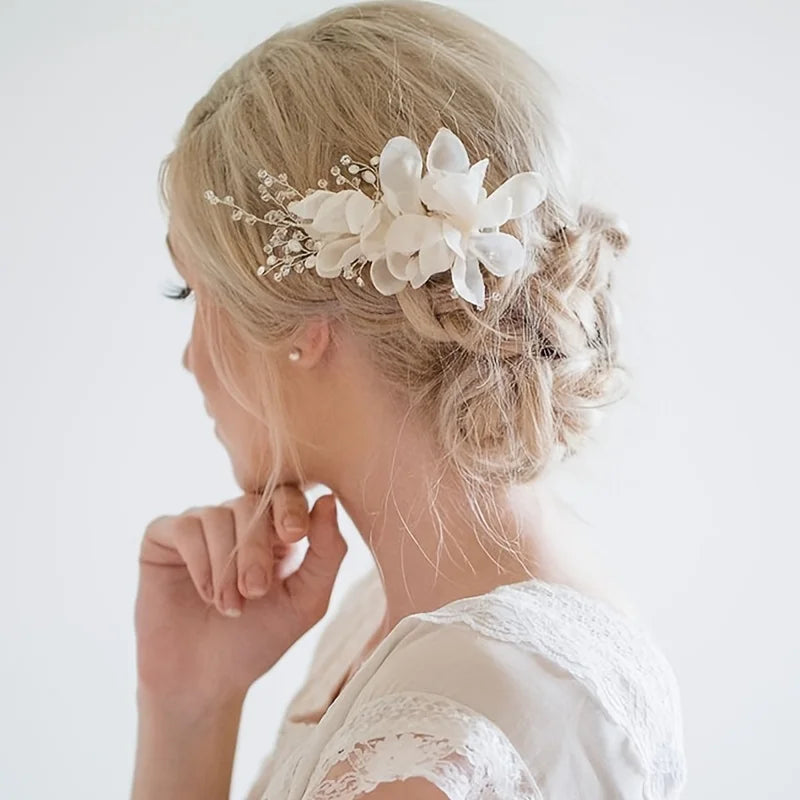 A woman with blonde hair styled in an elegant updo, adorned with a white floral hairpiece and delicate beads, wearing a white lace-trimmed top, looking thoughtfully to the side.