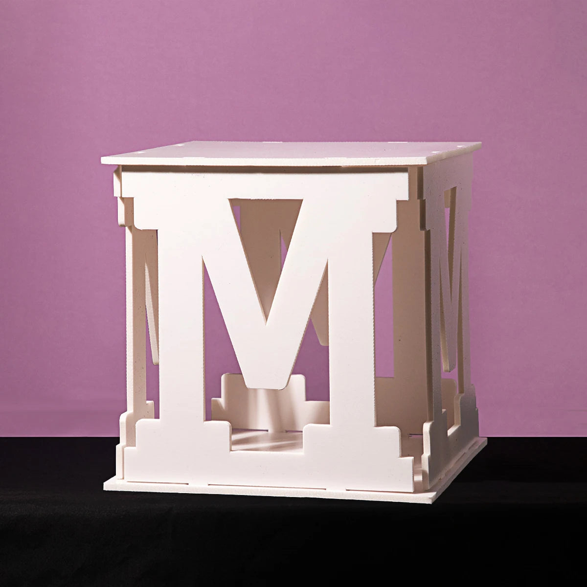White cubic structure with large cutout letters "M" on each side, set against a purple background. The design reveals a playful artistic style.