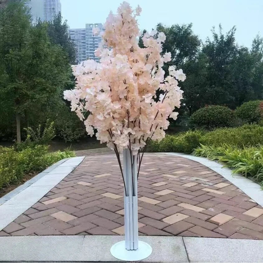 Artificial tree with light pink blossoms on a paved pathway in a park. Surrounded by greenery, with tall buildings faintly visible in the background.