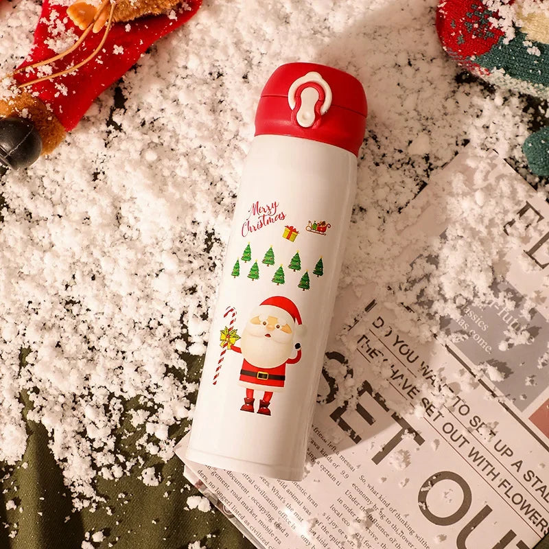 A festive white thermos with a red lid, featuring Santa, Christmas trees, gifts, and candy. It rests on a snow-dusted surface, surrounded by holiday decorations.