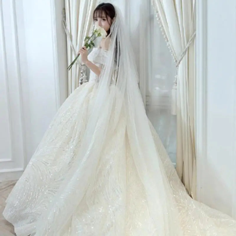 A bride in an elegant off-the-shoulder wedding gown with a long, flowing veil stands by white curtains, holding a bouquet of white flowers, looking serene and graceful.