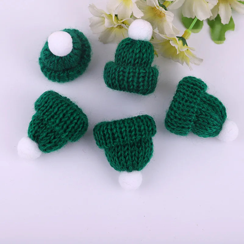 Five small, knitted green hats with white pom-poms are arranged on a white background near white flowers.