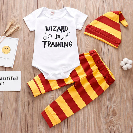 Baby outfit includes a white onesie with "Wizard in Training" text, red and yellow striped pants, and a matching hat, set on a wooden surface with decorative elements.