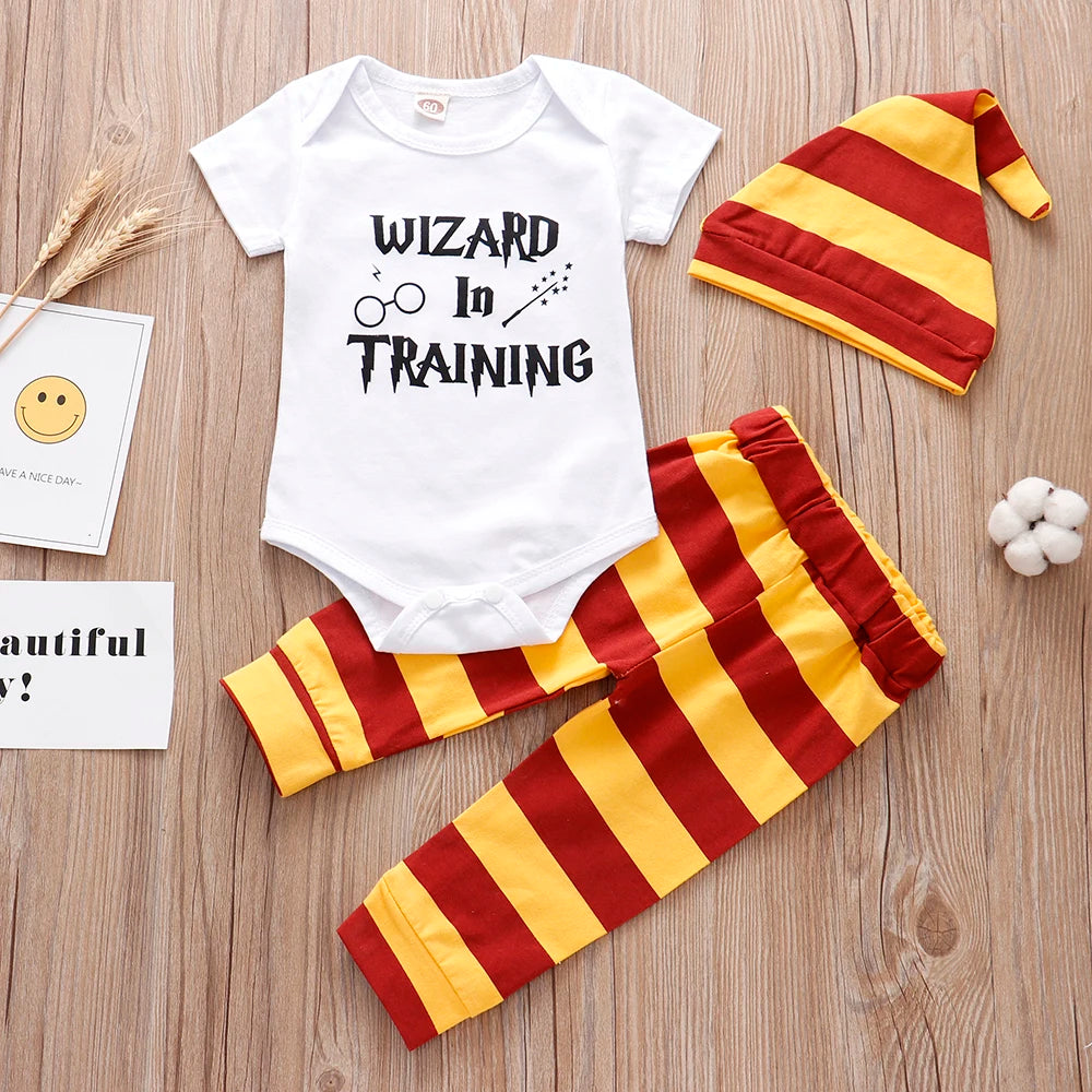 Baby outfit with a white onesie saying “Wizard in Training” and a yellow-red striped hat and pants on wooden floor. Nearby are a smiley card and cotton decoration.