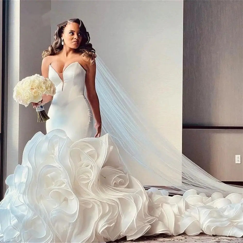 Bride in a sleek white wedding dress with elaborate ruffled train and long veil, holding a bouquet of white roses, looks to the side against a modern indoor backdrop.