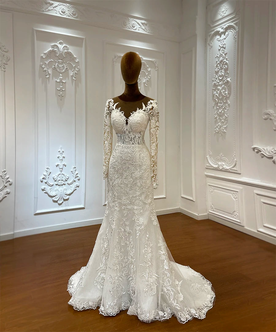A long-sleeved, lace wedding dress on a mannequin, featuring intricate floral patterns and a fitted silhouette, set in an ornate room with decorative white wall panels.