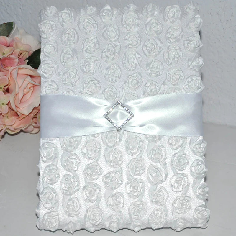 Elegant white textured wedding guest book adorned with small rosettes, a satin ribbon, and a diamond-shaped embellishment, placed next to pink roses.