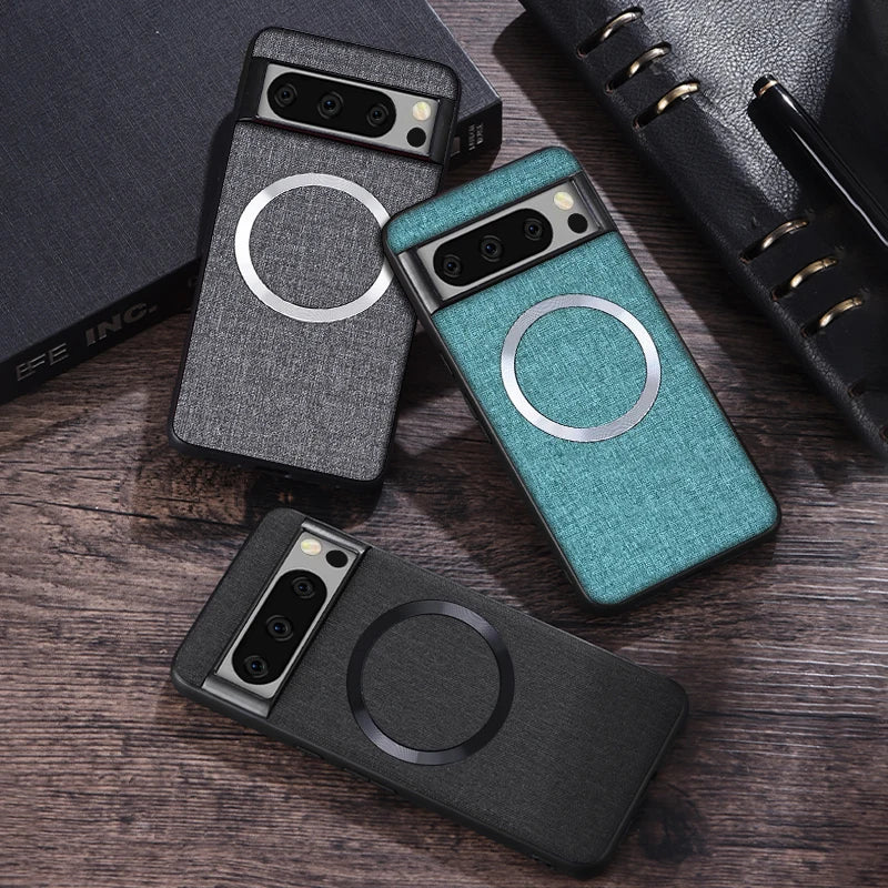 Three smartphone cases with a fabric-like texture and circular metal accents in black, gray, and teal are arranged on a wooden surface, accompanied by a black notebook.