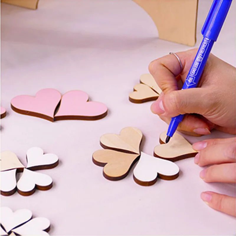A person decorates wooden heart shapes with a blue pen on a table. Pink, white, and natural wood-colored heart pieces are arranged neatly in groups.