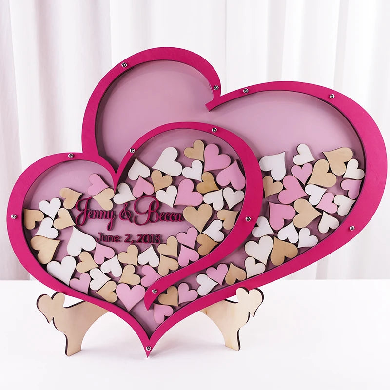 Pink heart-shaped shadow box with small pink, white, and beige hearts. Inscription reads "Jenny & Becca June 2, 2018." Decor piece with romantic theme, placed on white background.