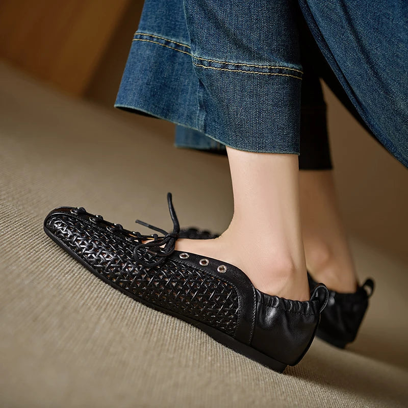 Woman wearing dark woven leather shoes paired with loose-fitting jeans, standing on a carpeted floor. The shoes feature lace-up detailing and metallic eyelets.