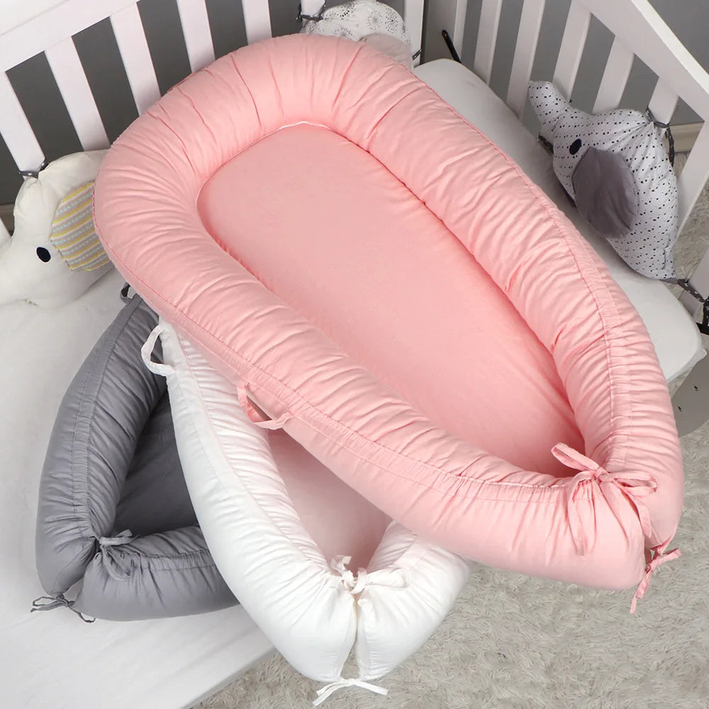Three baby nest cushions in pink, white, and gray are stacked in a crib. The cushions feature soft, padded edges and tied ends, providing a cozy spot for infants.