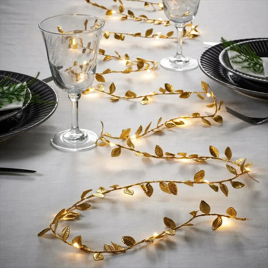 A table set elegantly with sparkling gold leaf garland lights, black plates with greenery, and clear wine glasses on a white tablecloth, creating a festive ambiance.