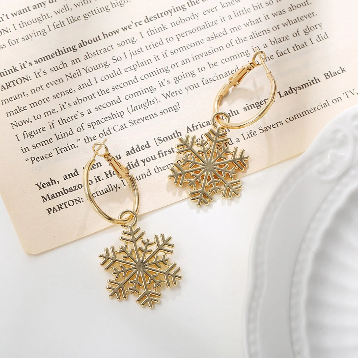Gold snowflake earrings on an open book page, with text partially visible. A scalloped edge of a white plate is on the right, adding elegance to the composition.