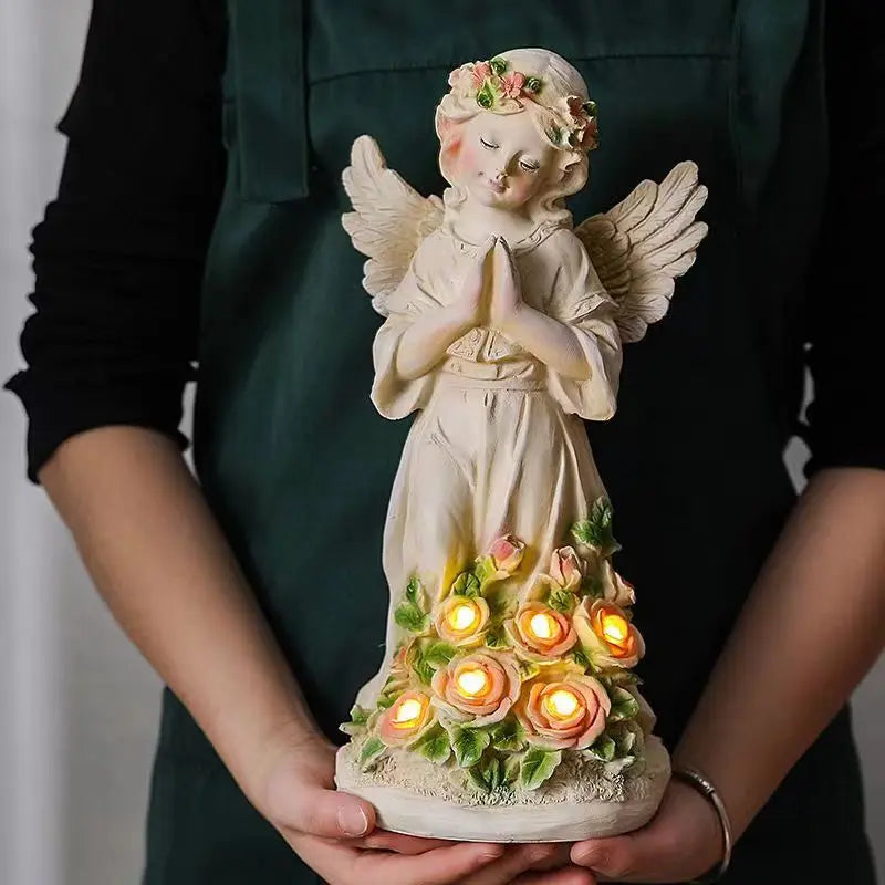 A person holds a statue of an angel with closed eyes and a floral crown, praying. The base is adorned with glowing roses, held delicately in both hands.