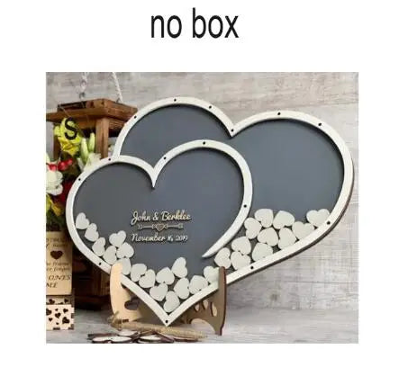 Heart-shaped guestbook frame with scattered wooden hearts, personalized with names and a date. Background includes a wooden crate and flowers. "No box" text at the top.